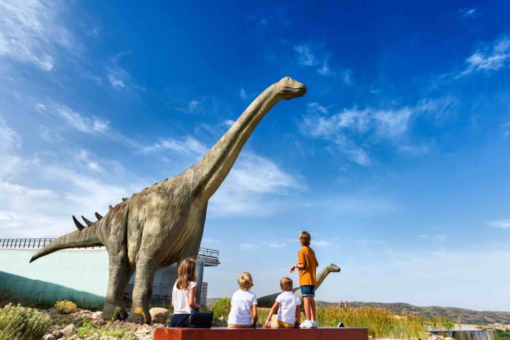 Cuatro niños contemplan una gran estatua de un dinosaurio al aire libre en un día soleado, con el cielo azul y las nubes de fondo.
