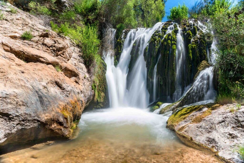 Una pintoresca cascada cae sobre rocas cubiertas de musgo hasta desembocar en una poza cristalina, rodeada de exuberante vegetación y rocas escarpadas, bajo un cielo azul brillante.