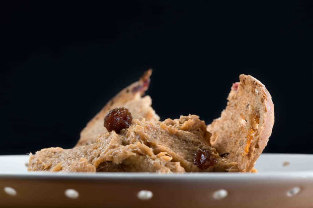 Un primer plano de un plato con una pasta para untar de color marrón, posiblemente paté o una salsa para mojar pan, adornada con pasas y acompañada de dos rebanadas de pan sobre un fondo negro.