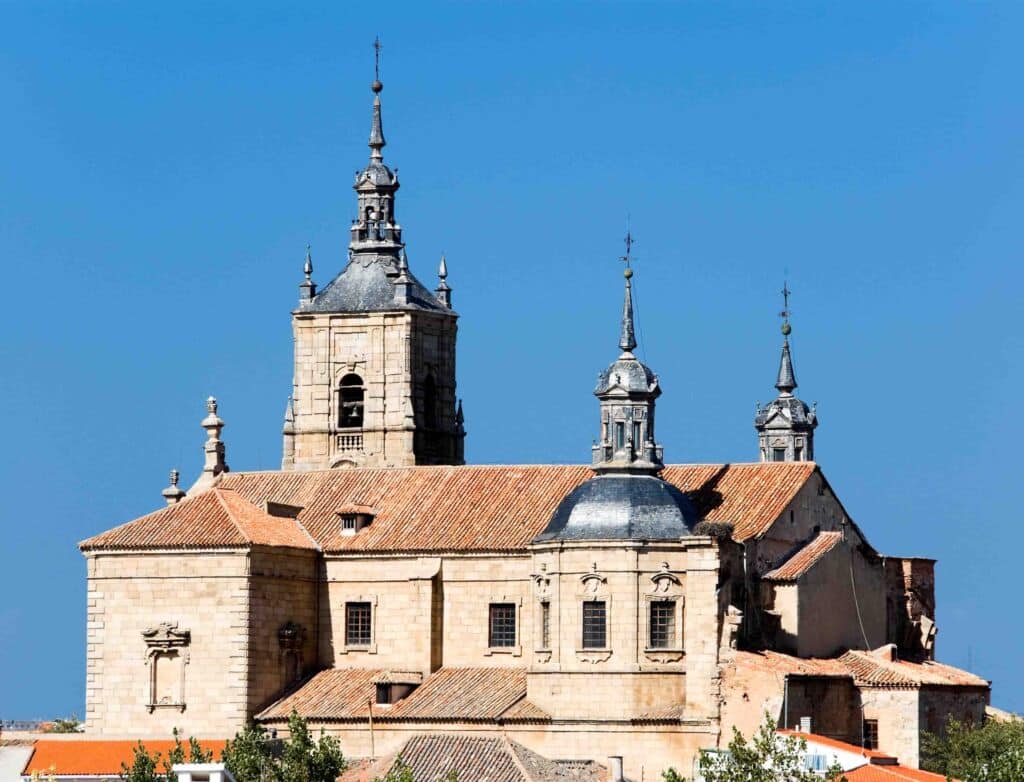 Iglesia histórica de piedra con tres altas agujas y un tejado de tejas rojas que se recorta contra un cielo azul claro, enmarcada parcialmente por las copas verdes de los árboles en la parte inferior de la imagen.