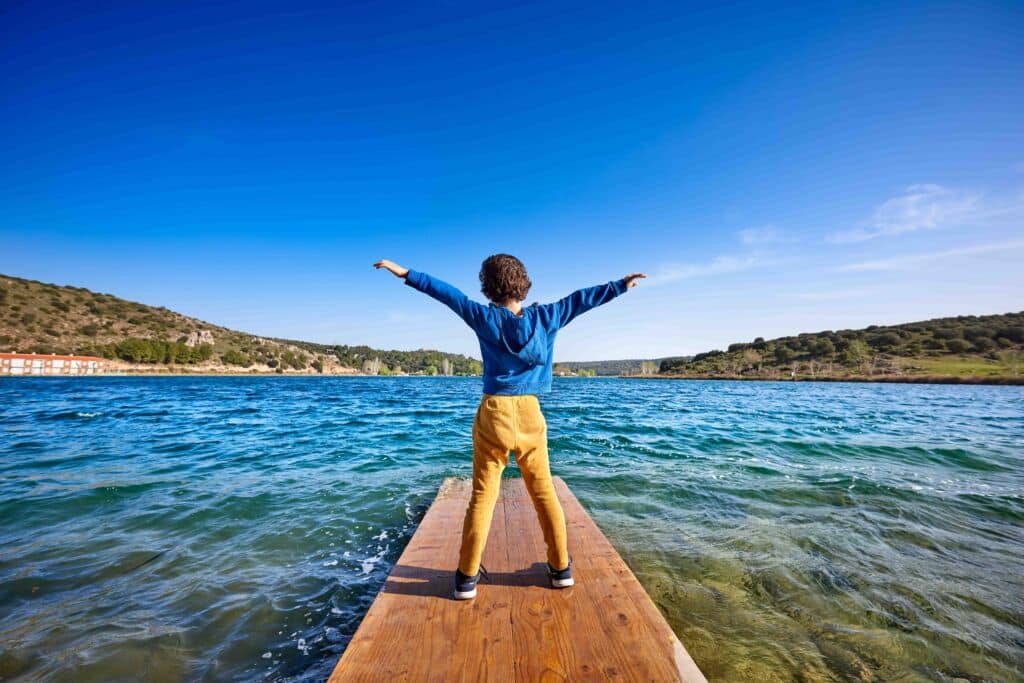 Un niño está de pie al final de un embarcadero de madera, con los brazos extendidos, mirando hacia un lago azul rodeado de colinas y vegetación bajo un cielo despejado.