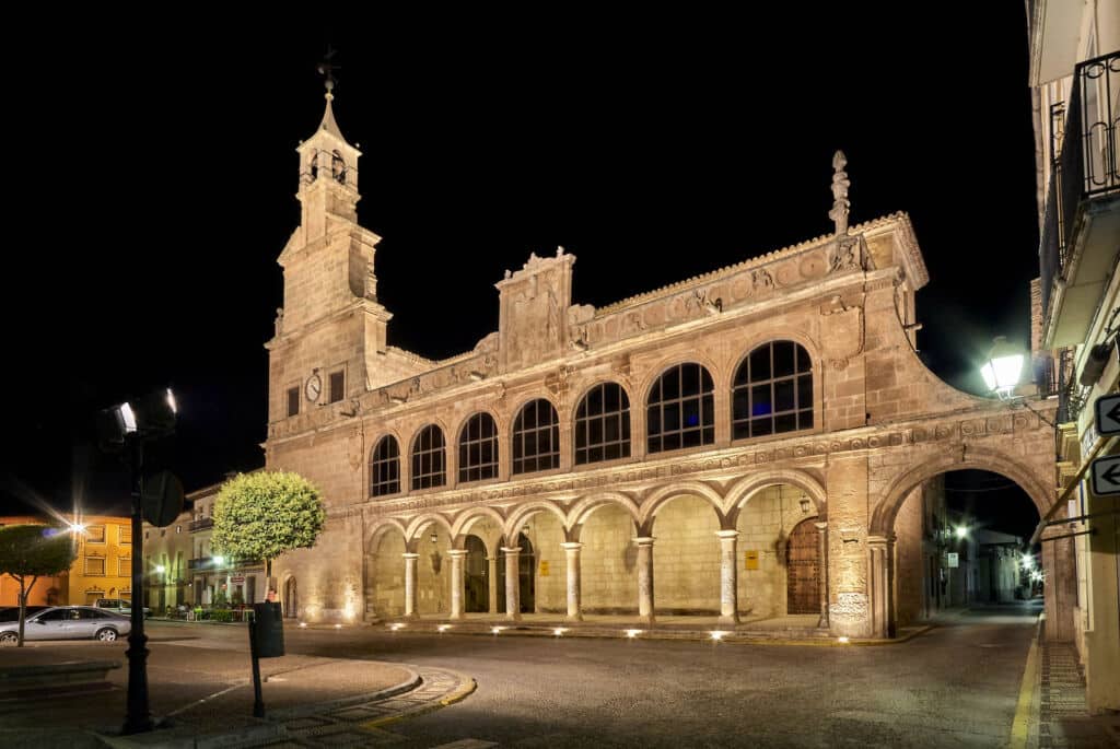 Edificio histórico de piedra con columnas arqueadas y grandes ventanales, iluminado por la noche. Un alto campanario se eleva a la izquierda, y las farolas proyectan una luz cálida sobre la plaza tranquila y desierta que hay frente a él.