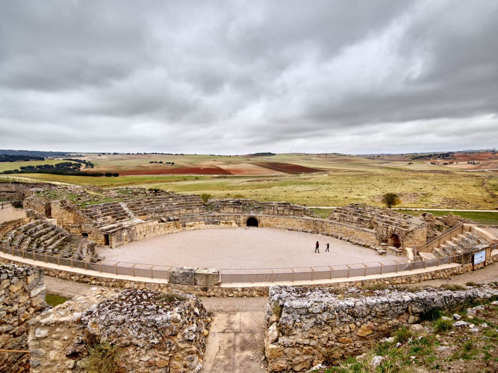 Ruinas de un antiguo anfiteatro romano con gradas de piedra, rodeadas de campo abierto bajo un cielo nublado. Dos pequeñas figuras caminan por el centro de la arena.