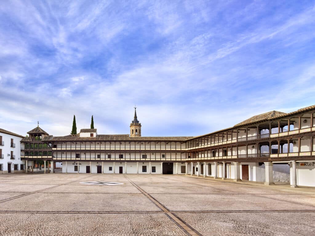 Un patio amplio y vacío, con suelo de adoquines, rodeado por un edificio de dos plantas con balcones de madera. Al fondo, bajo un cielo azul con nubes dispersas, se alzan una torre con aguja y dos árboles altos.