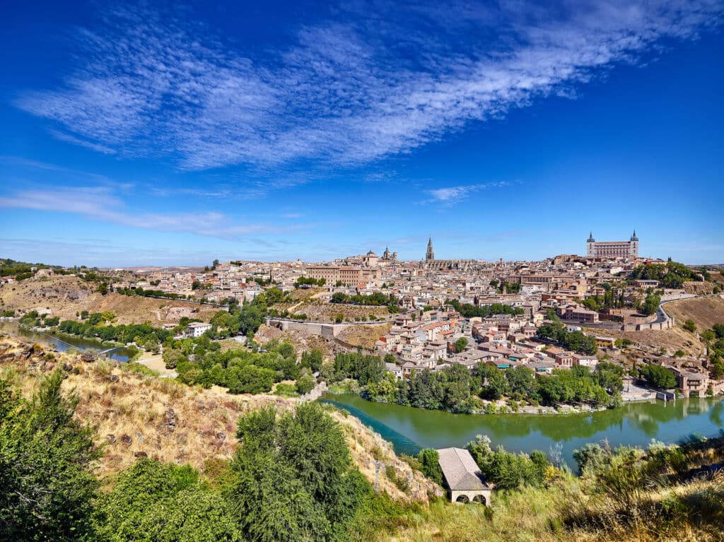 Una vista panorámica de una ciudad histórica con un río que serpentea entre verdes colinas, edificios de piedra y torres imponentes, bajo un cielo azul brillante salpicado de nubes tenues.