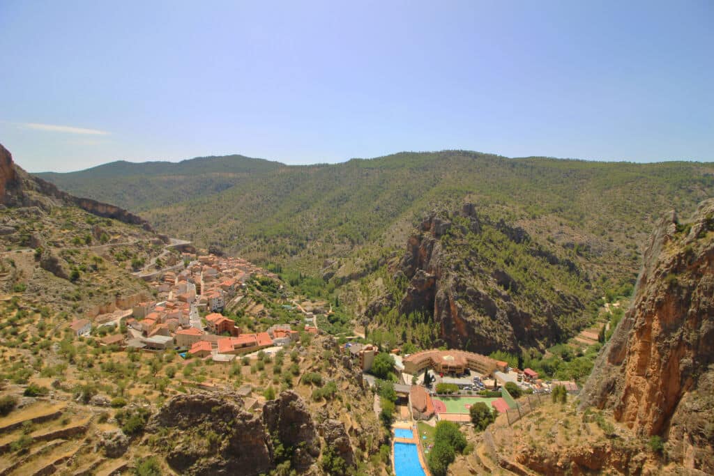 Un pueblo con tejados de terracota se encuentra enclavado en un valle verde, rodeado de colinas rocosas y bosques. En primer plano, bajo un cielo despejado, se divisa una piscina de un azul intenso.