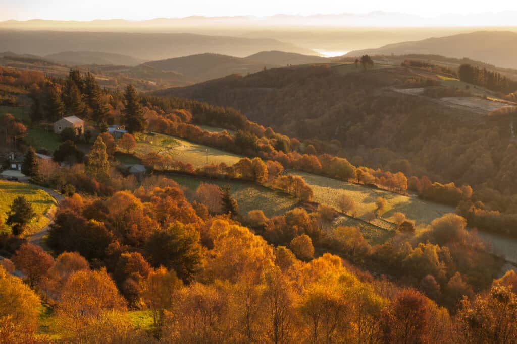 Colinas onduladas cubiertas de coloridos árboles otoñales, casas dispersas y campos bajo una puesta de sol dorada, con montañas y una masa de agua lejana visibles al fondo.