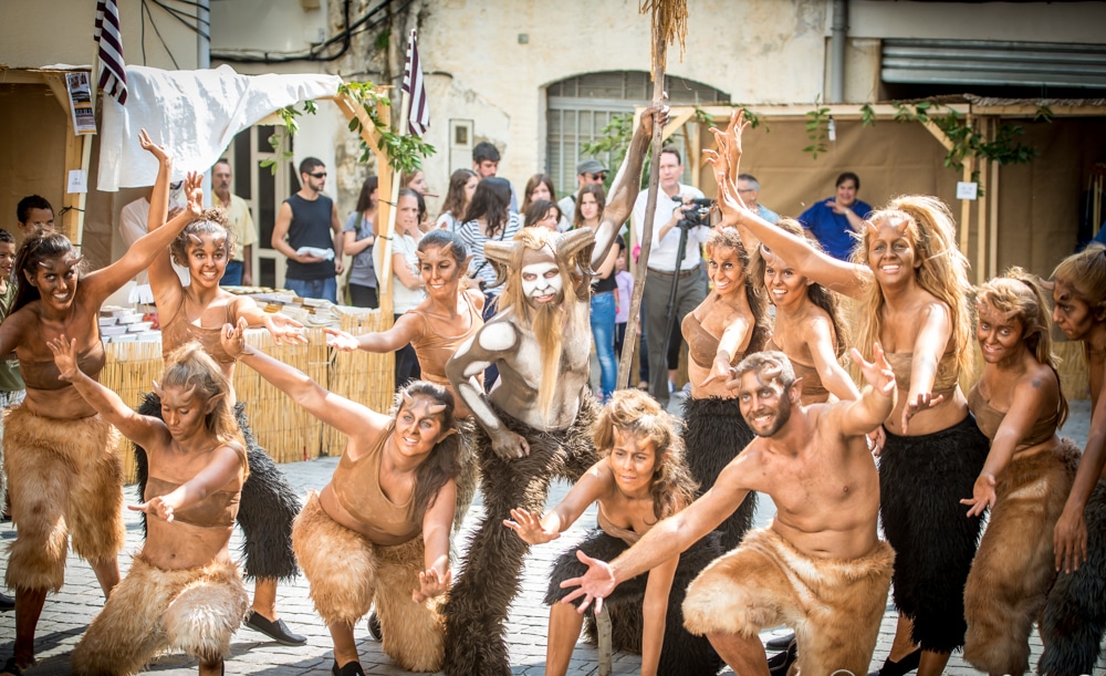Un grupo de artistas vestidos como criaturas míticas, con trajes peludos y las caras pintadas, posan con energía al aire libre en un animado festival, mientras la gente observa al fondo.