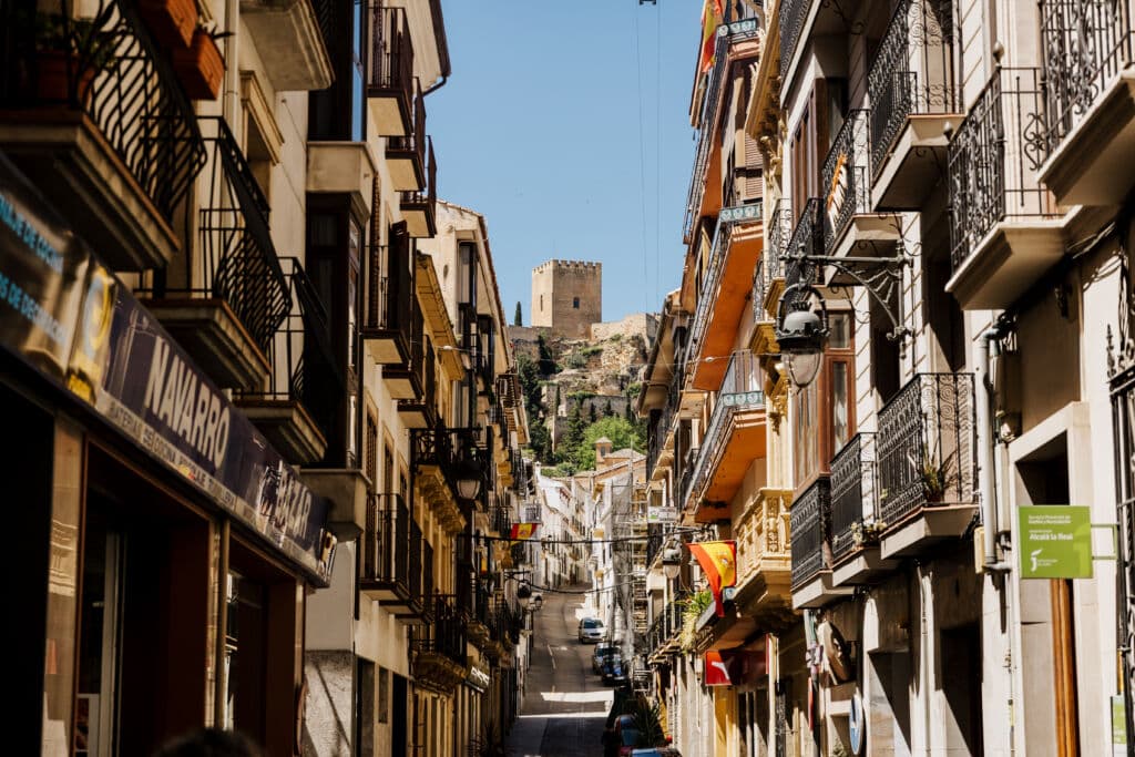 Una calle estrecha, flanqueada por balcones y tiendas, asciende hacia un antiguo castillo de piedra bajo un cielo azul y despejado. De algunos edificios cuelgan banderas españolas y hay unos cuantos coches aparcados a lo largo de la calle.