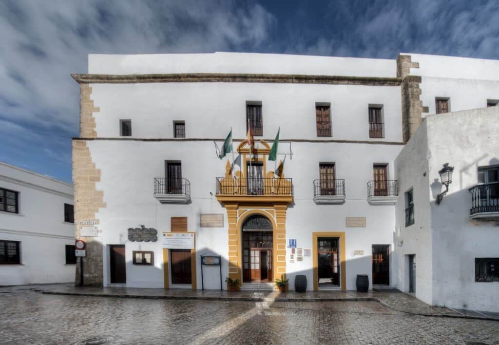 Edificio blanco con una entrada en arco con ribetes amarillos, tres balcones y varias banderas verdes y amarillas. Calle empedrada y cielo nublado al fondo.