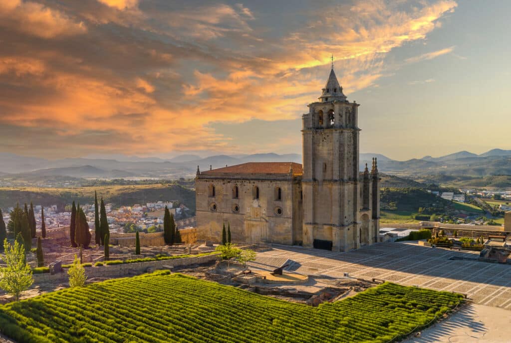 Una histórica iglesia de piedra con un alto campanario se alza junto a unos cuidados jardines al atardecer, con las montañas y un pequeño pueblo visibles al fondo bajo un espectacular cielo anaranjado.