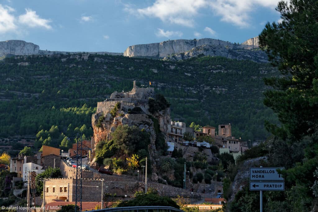 Un pueblo con edificios de piedra se alza sobre una colina rocosa, con altas montañas al fondo. En primer plano se ve una carretera sinuosa y señales que indican Vandellòs, Mora y Pradip. El cielo está parcialmente nublado.