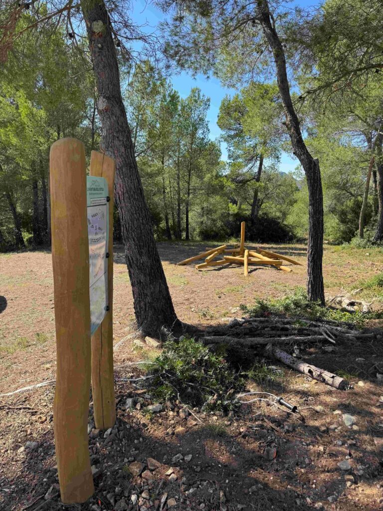 Junto a unos árboles y a un claro de tierra, con una estructura de madera para trepar al fondo, se alza un cartel informativo de madera, rodeado por el bosque y la luz del sol.