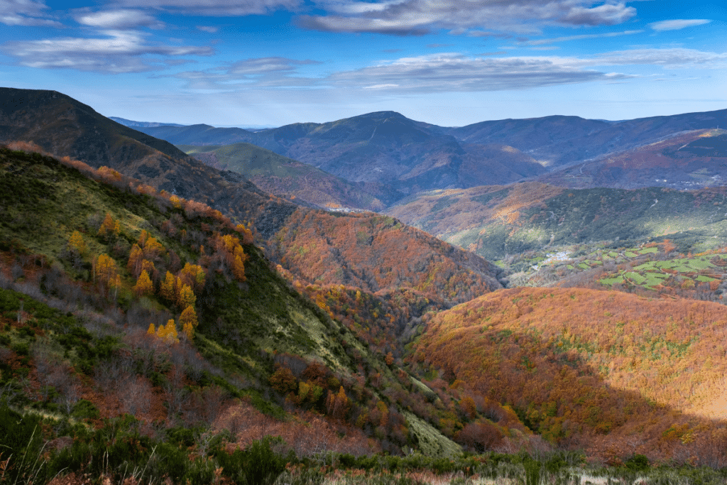 Un pintoresco valle montañoso con árboles otoñales en tonos naranjas, amarillos y verdes bajo un cielo azul salpicado de nubes. La luz del sol ilumina algunas partes de las colinas onduladas y las montañas lejanas.