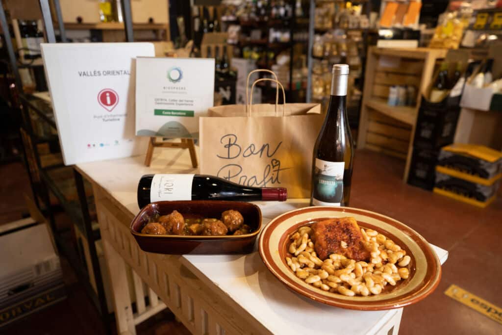En el mostrador de una tienda, con estanterías llenas de productos envasados al fondo, se ven dos platos con comida, dos botellas de vino y una bolsa de papel marrón con la etiqueta «Bon Profit».