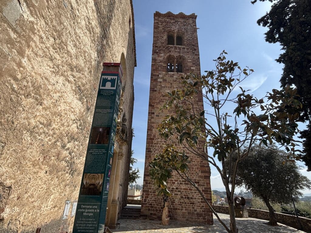 Junto a un antiguo edificio de piedra, con un cartel informativo y árboles frondosos a su alrededor, se alza un campanario alto y rectangular de ladrillo, bajo un cielo azul y despejado.