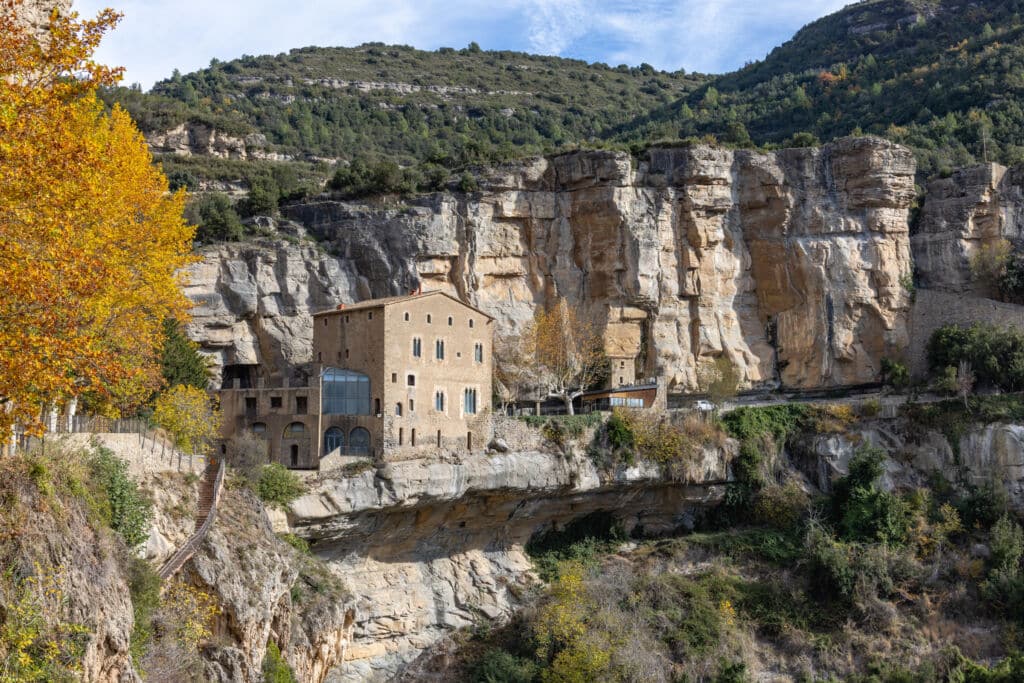 Un edificio de piedra se alza encajado en unos acantilados rocosos, rodeado de árboles y follaje otoñal, con las montañas y un cielo despejado al fondo.