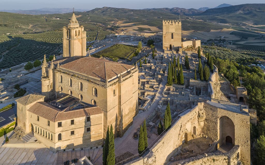 Vista aérea de una antigua fortaleza y un complejo eclesiástico rodeados de árboles, ruinas y colinas onduladas bajo un cielo despejado, con olivares al fondo.