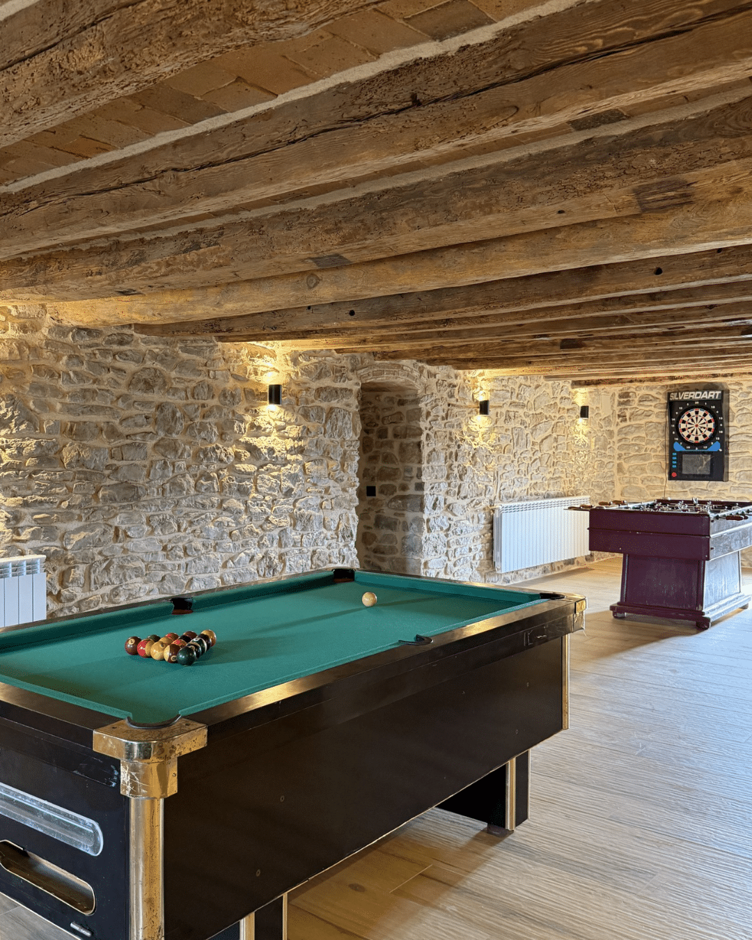Una mesa de billar con un triángulo de bolas en primer plano y un futbolín y una diana electrónica al fondo, en una habitación con paredes de piedra, durante un verano rural en la Comunitat Valenciana.