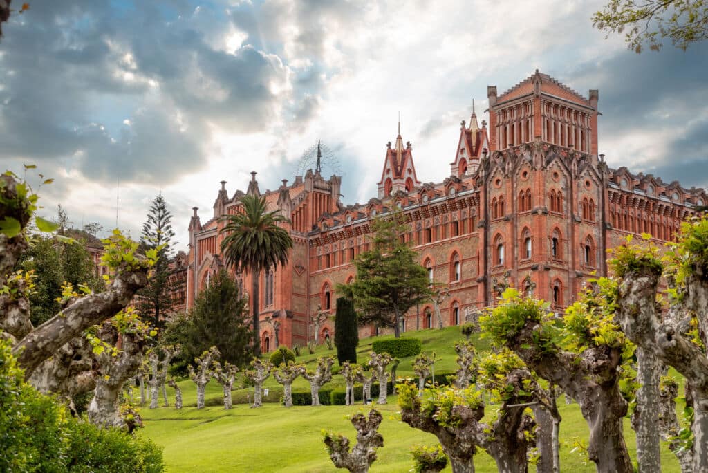 Un edificio grande y ornamentado de ladrillo rojo, con arcos apuntados y torres, situado detrás de un césped verde y bien cuidado, con árboles podados.