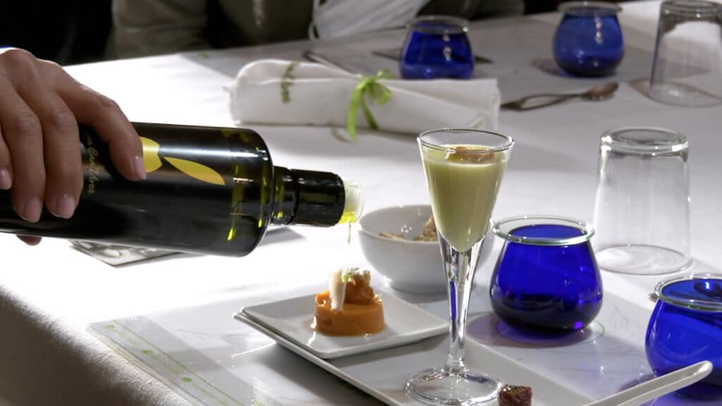 Una mano vierte aceite de oliva sobre un pequeño aperitivo colocado sobre una mesa blanca, en la que hay un vaso alto y cristalería azul.