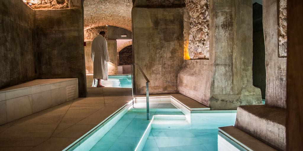 Una persona vestida con una túnica blanca se encuentra junto a una piscina termal cubierta, rodeada de muros y arcos de piedra de época.