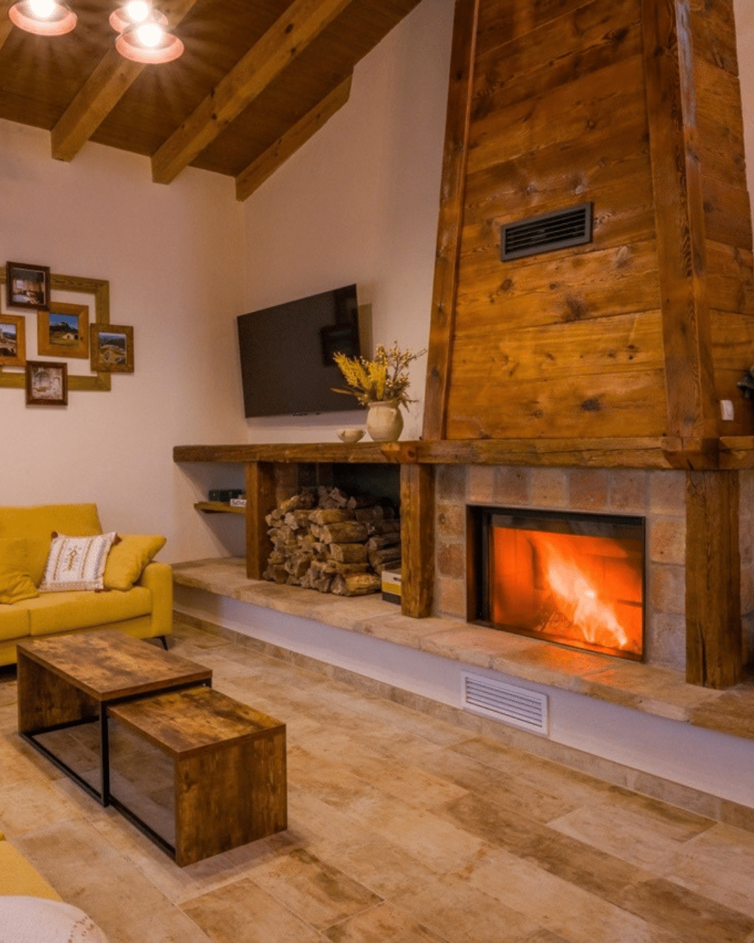 En el salón hay un sofá amarillo, junto a una chimenea de piedra con repisa de madera, leña y un televisor colgado en la pared, lo que evoca un verano rural en la Comunitat Valenciana.