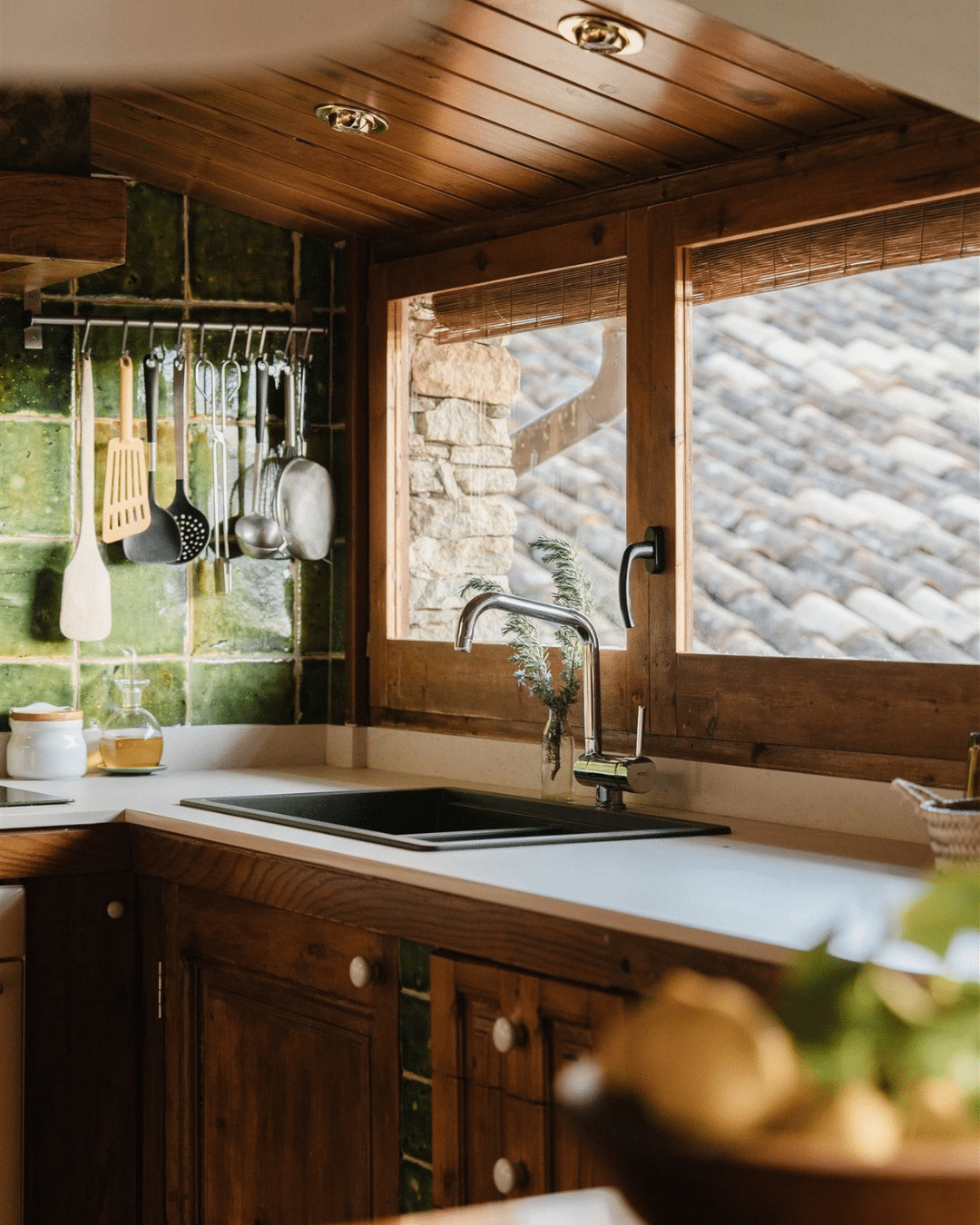 Grifo de cocina cromado empotrado en una encimera frente a una ventana, con utensilios de cocina colgados de una pared de azulejos verdes, que evoca un verano rural en la Comunidad Valenciana.