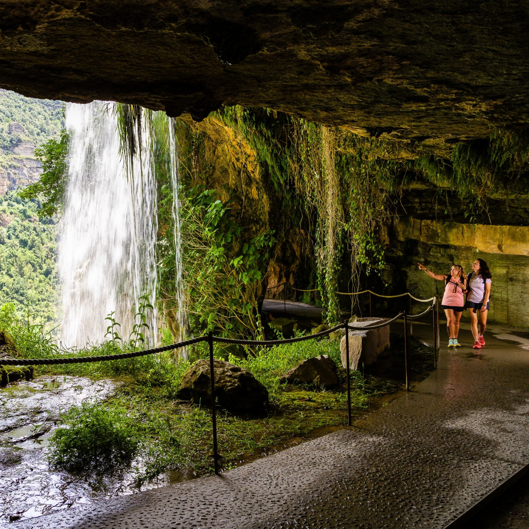 Dos excursionistas caminan por un sendero detrás de una cascada durante un verano en el campo de la Comunitat Valenciana; una de las mujeres señala hacia el agua que cae en cascada.