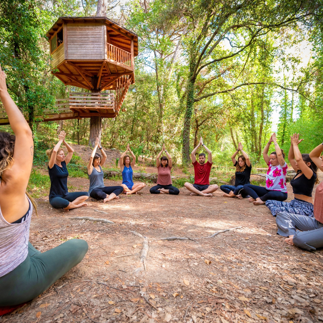 Un grupo de personas está sentado en círculo en el suelo del bosque realizando una postura de yoga, disfrutando de un verano en el campo en la Comunitat Valenciana, con una casa del árbol de madera al fondo.