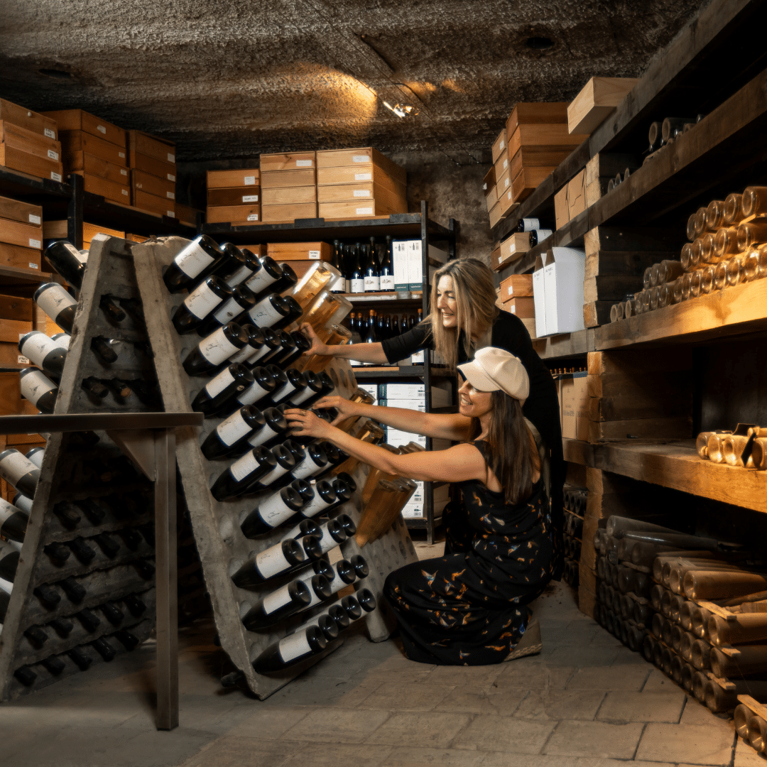 Dos mujeres en una bodega, durante un verano en el campo de la Comunidad Valenciana, dando la vuelta a las botellas almacenadas en una estantería inclinada con estructura en forma de A.