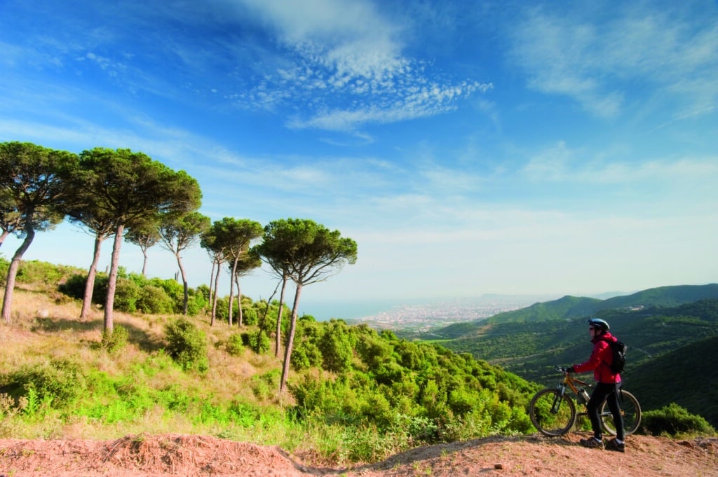 Un ciclista de montaña con una chaqueta roja está de pie en una cresta desde la que se divisa una ciudad costera.