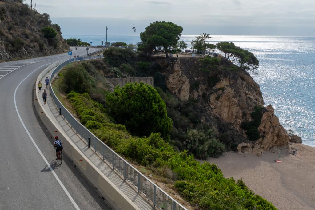 Un ciclista pedalea por una carretera costera junto a un acantilado con vistas a una playa de arena y al mar.
