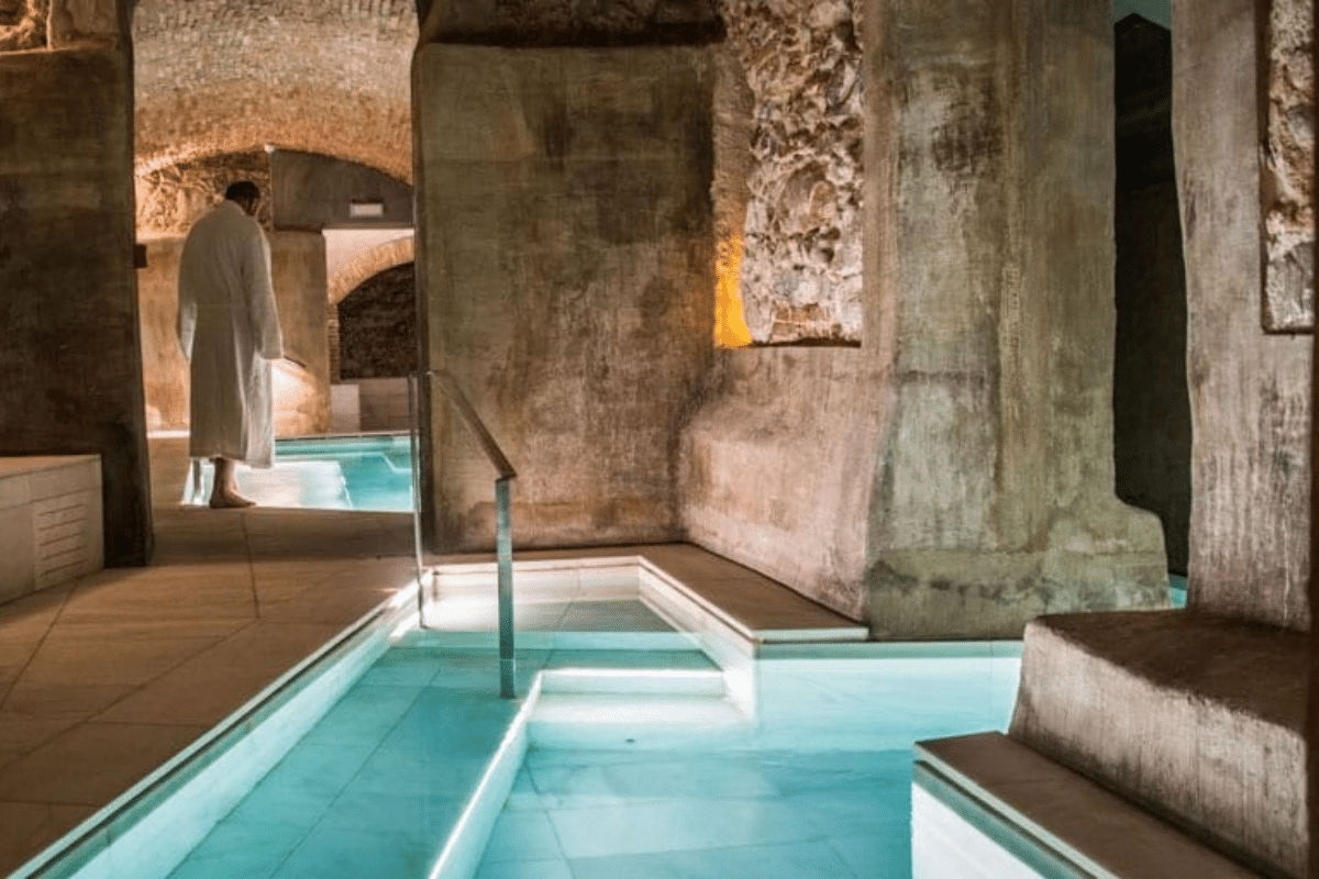 Una persona vestida con albornoz se encuentra de pie al borde de una piscina termal de color turquesa, en el interior de una caverna con paredes de piedra, lo que evoca un verano rural en la Comunidad Valenciana.
