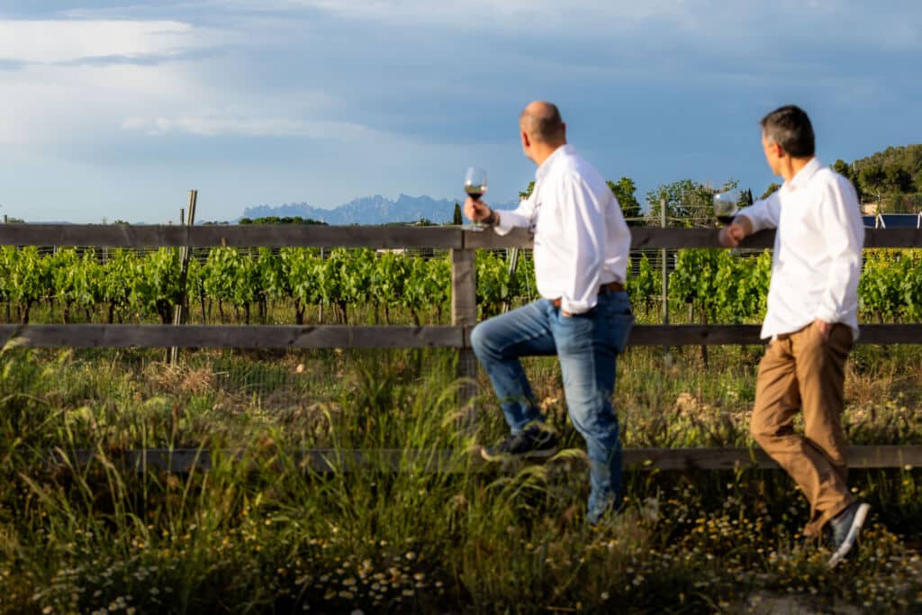 Dos hombres con camisa blanca, con una copa de vino en la mano, están apoyados contra una valla de madera con vistas a un viñedo.