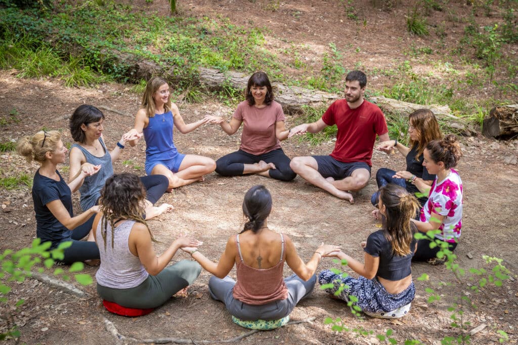 Un grupo variado de nueve personas está sentado en círculo en el suelo, en medio de un bosque, cogidas de la mano con las palmas hacia arriba.